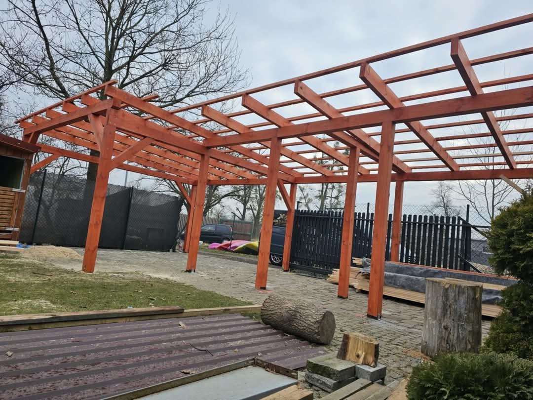 Konstrukcja altany z impregnowanego drewna w trakcie montażu, widoczne elementy więźby dachowej, w tle ogrodzenie posesji i drzewa bez liści.