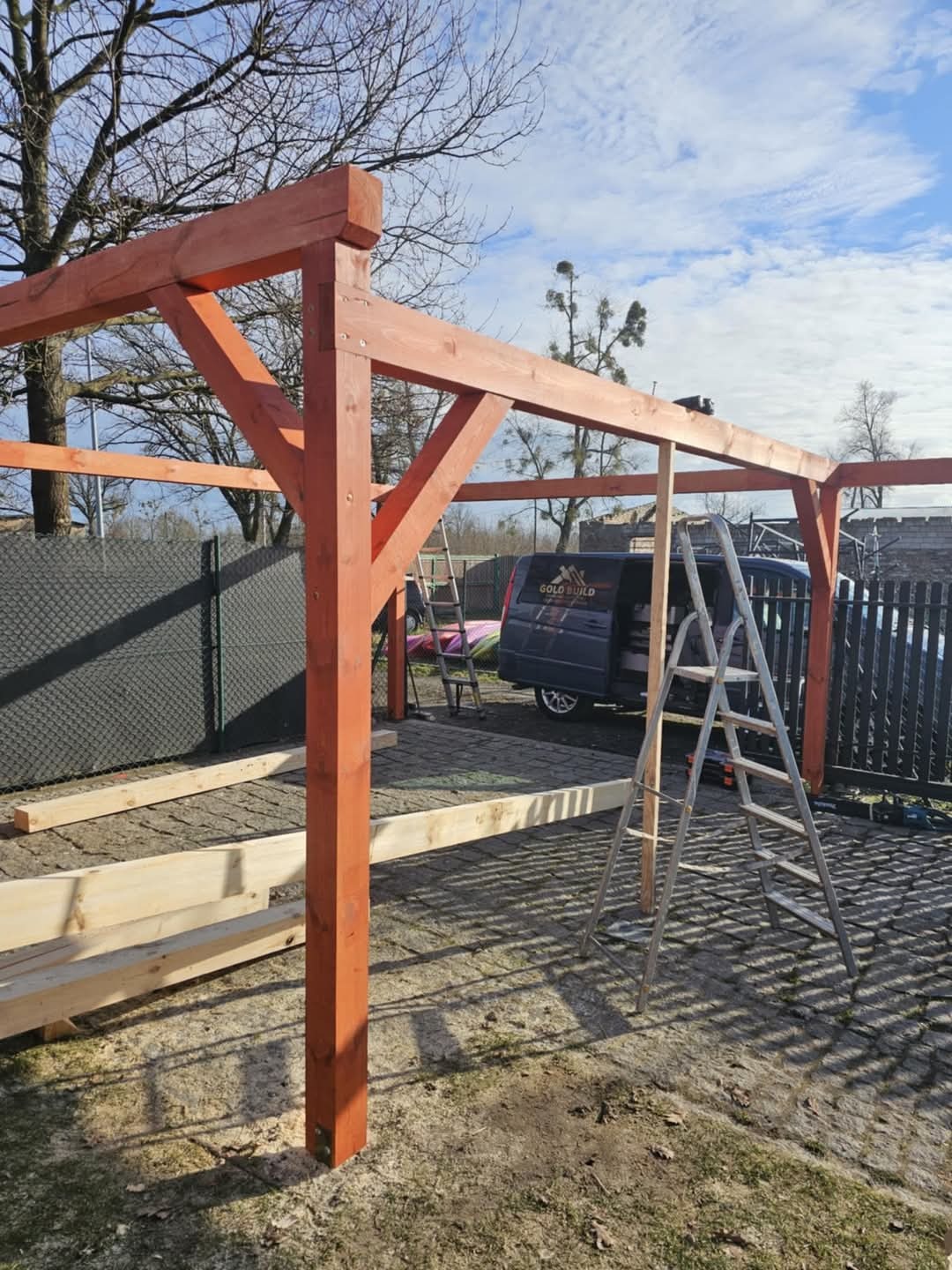 Konstrukcja drewniana altany w trakcie montażu, widoczne słupy i elementy nośne zaimpregnowanego drewna, drabina oparta o konstrukcję, w tle samochód dostawczy z logo firmy.