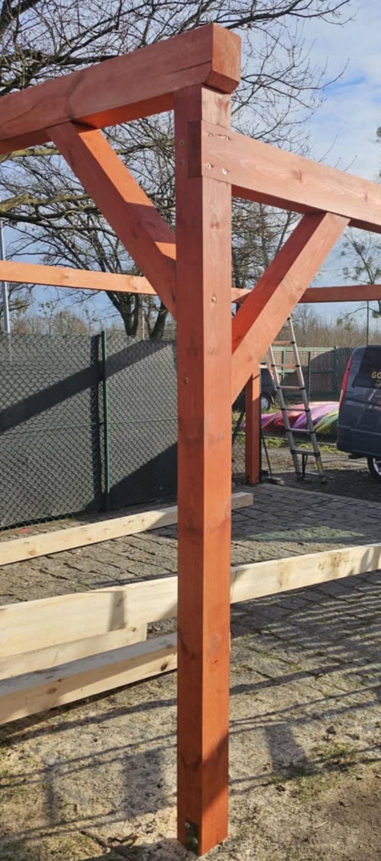 Konstrukcja drewniana altany w trakcie montażu, widoczne elementy nośne z drewna o pomarańczowym zabarwieniu, na tle ogrodzenia z siatki i częściowo widocznego samochodu dostawczego.