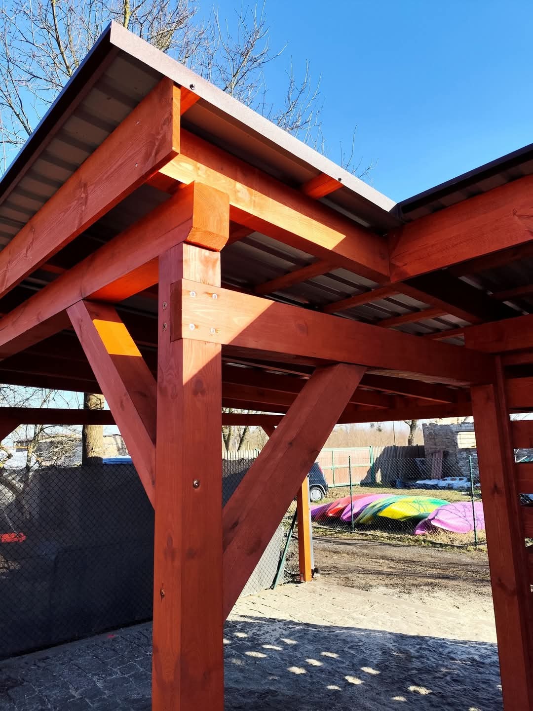 Konstrukcja drewniana z metalowym zadaszeniem, widok z dołu na połączenie belek i słupów, w tle kolorowe kajaki i ogrodzenie z siatki.
