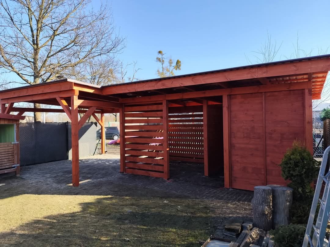 Drewniana konstrukcja zadaszona, częściowo zabudowana panelami z poziomych listew, pomalowana na czerwono-brązowy kolor, z widoczną konstrukcją dachu, na tle trawnika i drzew bez liści.