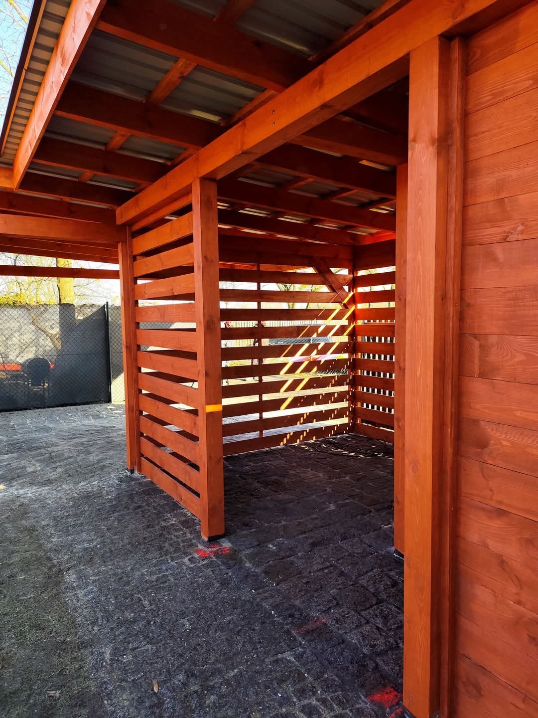 Nowo wybudowana, pomalowana na brązowo drewniana konstrukcja z metalowym dachem, z poziomymi listwami po bokach, stojąca na szarej kostce brukowej.