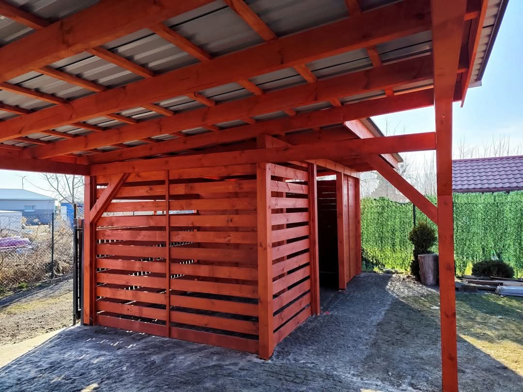 Drewniana konstrukcja z zadaszeniem z blachy trapezowej, pomalowana na pomarańczowo-czerwony kolor, z widocznymi poziomymi deskami tworzącymi ściany boczne, na tle zielonego żywopłotu i błękitnego...