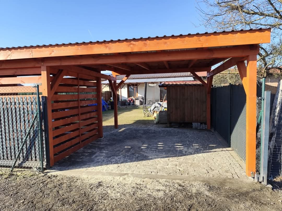 Drewniana wiata garażowa z zadaszeniem z blachy trapezowej, częściowo zabudowana poziomymi deskami, na posesji z kostką brukową i ogrodzeniem z siatki.