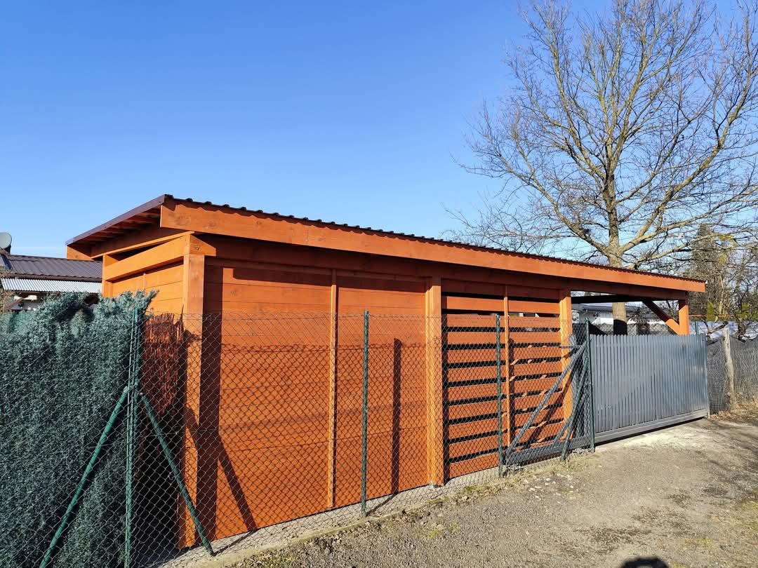 Drewniana konstrukcja z zadaszeniem z blachy falistej, widoczna zza ogrodzenia z siatki, na tle błękitnego nieba i drzewa bez liści.