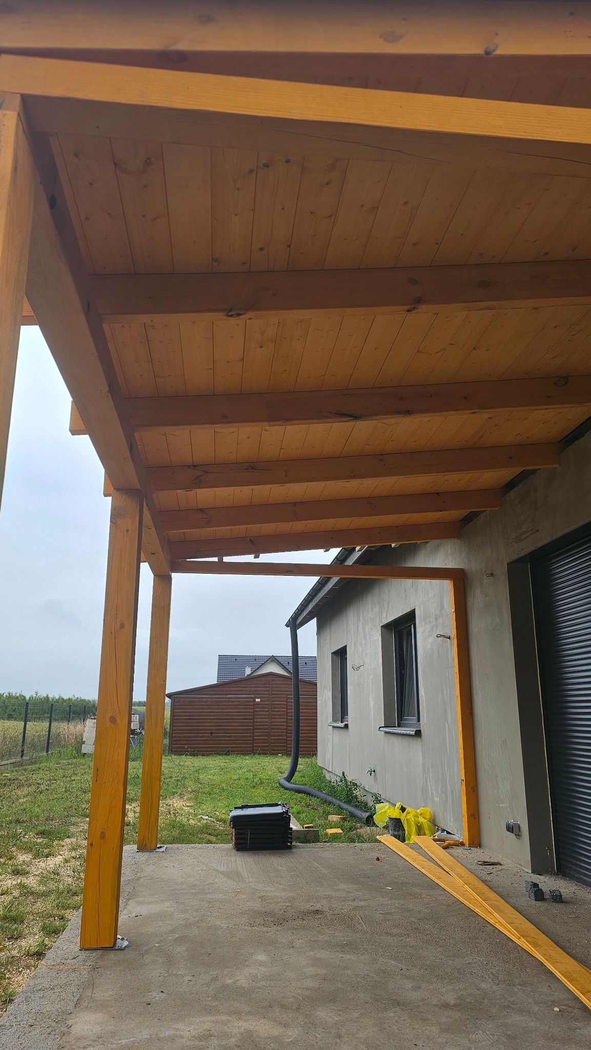 Drewniana konstrukcja zadaszenia tarasu w trakcie budowy, widoczne słupy i deski, tło stanowi budynek z szarą elewacją i zielony trawnik.