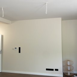 GIREM - Jasne wnętrze po remoncie: panele podłogowe w odcieniach brązu, świeżo pomalowane ściany w kolorze złamanej bieli, gniazdka i wyjścia na kable w ścianie, oraz ozdobna lampa.