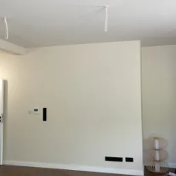 Jasne wnętrze po remoncie: panele podłogowe w odcieniach brązu, świeżo pomalowane ściany w kolorze złamanej bieli, gniazdka i wyjścia na kable w ścianie, oraz ozdobna lampa.