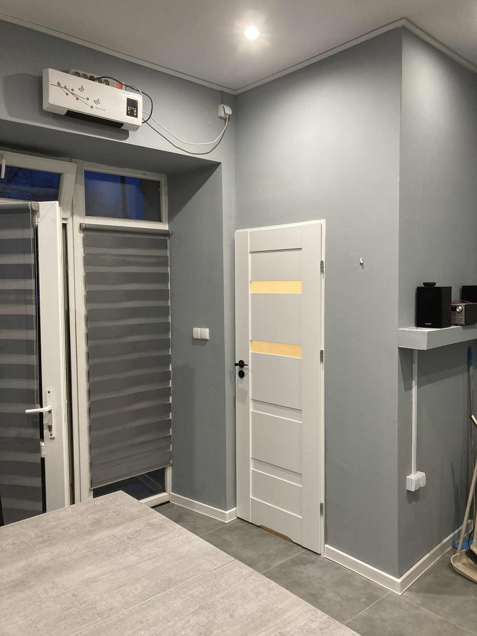 Szara ściana z białą stolarką drzwiową i oknem z żaluzjami. Na ścianie zamontowany grzejnik elektryczny i półka z głośnikami. Nowoczesne wykończenie.