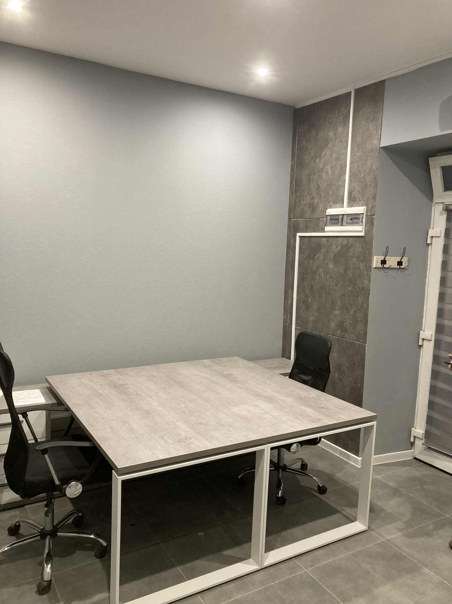 Nowoczesne biuro z szarymi ścianami, dużym stołem z blatem imitującym beton i czarnymi krzesłami obrotowymi. Minimalistyczne oświetlenie sufitowe.