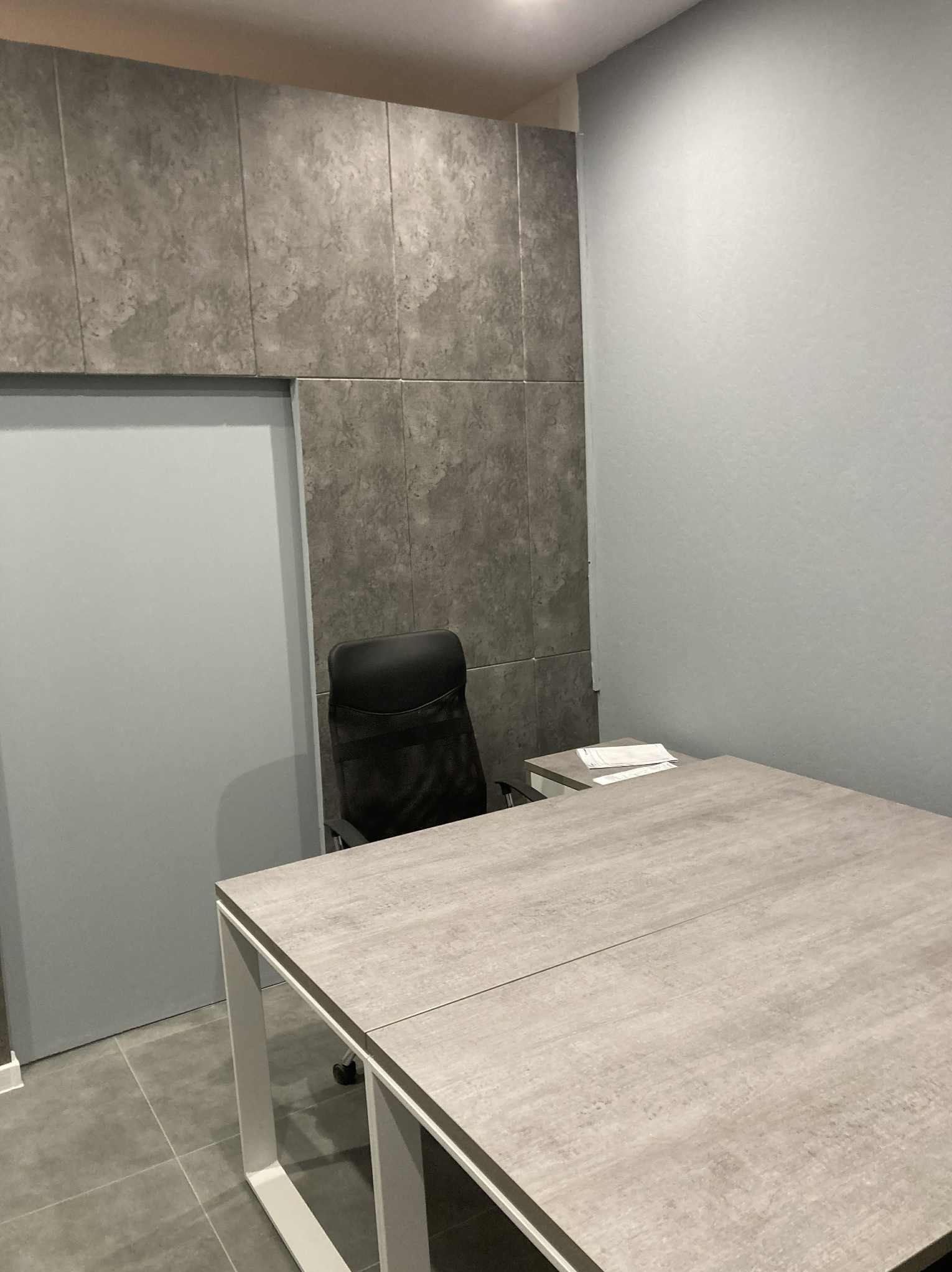 Wykończone biuro z szarymi panelami ściennymi, biurkiem i krzesłem. Minimalistyczny design, neutralna kolorystyka. Widok na fragment ściany i podłogi.