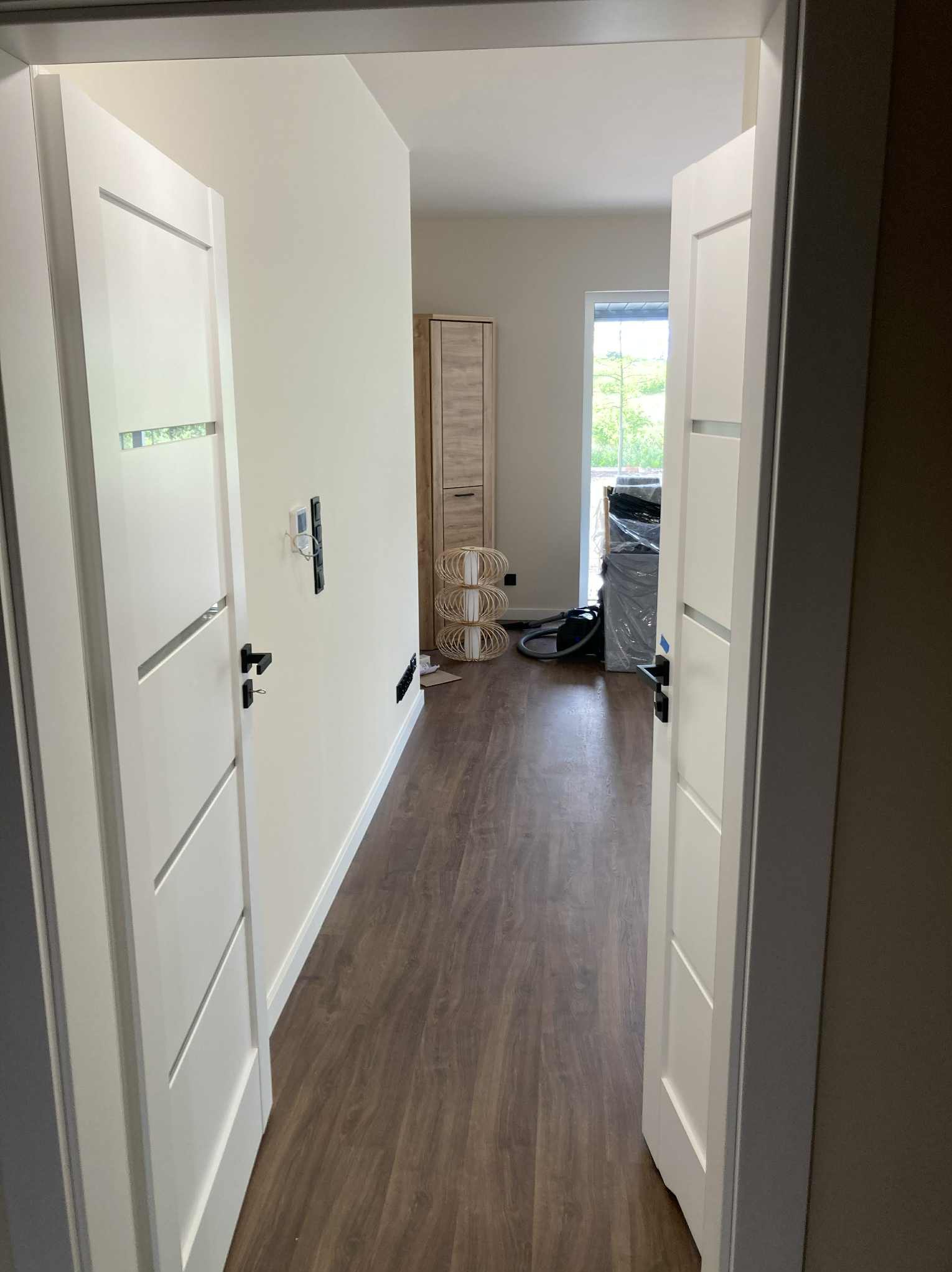 Nowoczesne, białe drzwi wewnętrzne z czarną klamką, zamontowane w jasnym wnętrzu z panelami podłogowymi. Widok z perspektywy przechodzącego przez próg.