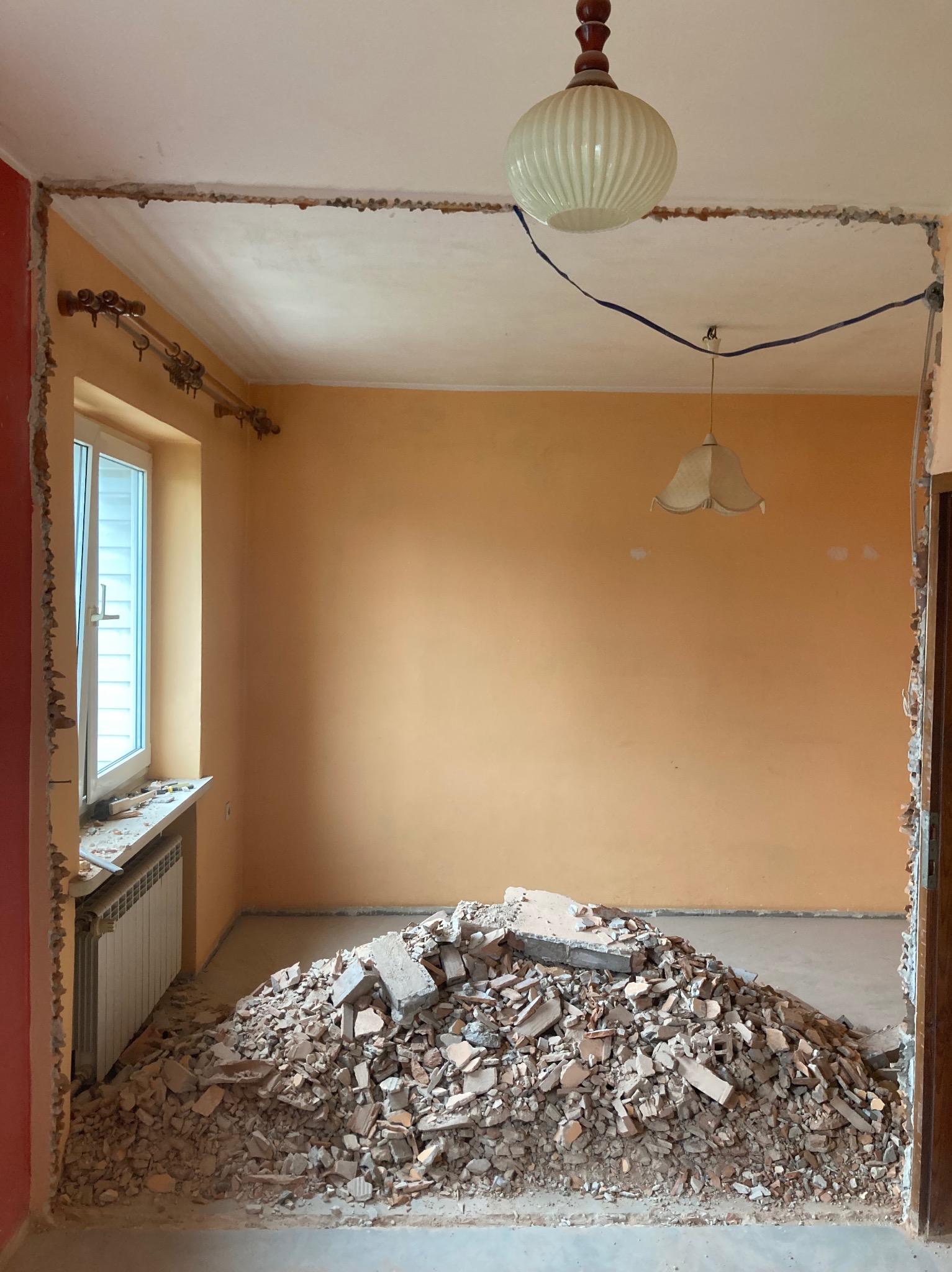 Pokój w trakcie demontażu: gruz na podłodze, ściany częściowo skute, widoczne instalacje elektryczne i grzejnik. Przygotowanie do prac remontowych.