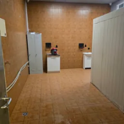 Wnętrze pomieszczenia wyłożonego brązowymi płytkami z widoczną szafką metalową, umywalkami, oraz sprzętem czyszczącym. Na suficie widoczne kamery monitoringu.