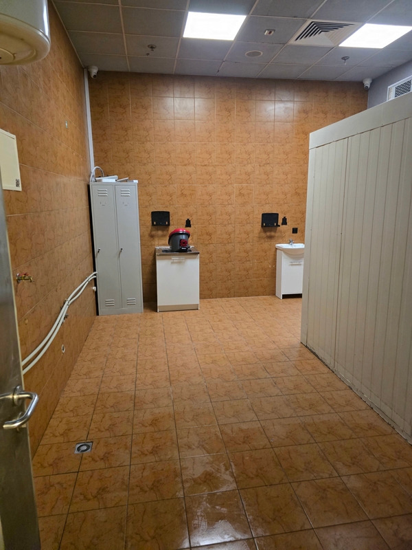 Wnętrze pomieszczenia wyłożonego brązowymi płytkami z widoczną szafką metalową, umywalkami, oraz sprzętem czyszczącym. Na suficie widoczne kamery monitoringu.