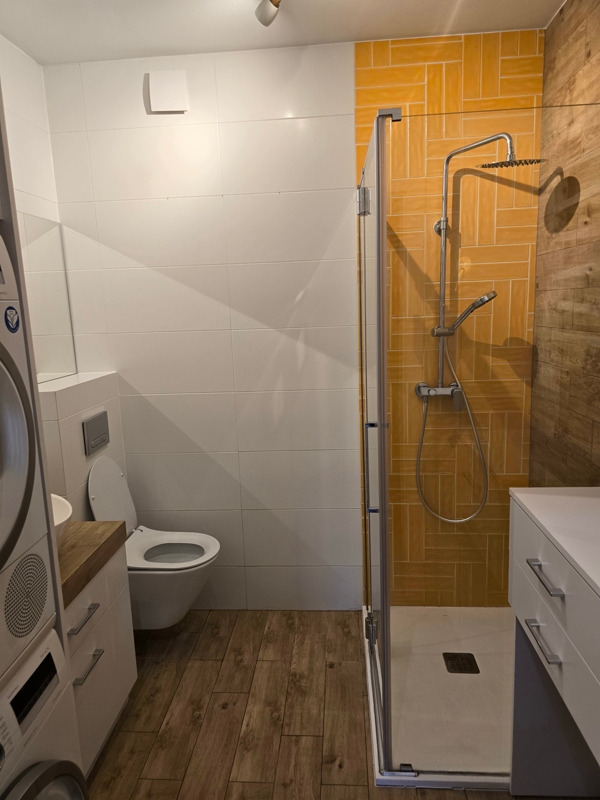 Nowoczesna łazienka z kabiną prysznicową typu walk-in, toaletą wiszącą, pralką i suszarką w zabudowie, oraz żółtymi płytkami ułożonymi w jodełkę w strefie prysznica.