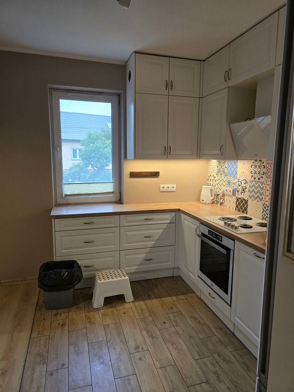 Widok na jasną, narożną kuchnię z drewnianą podłogą, białymi szafkami i blatem, z widocznym koszem na śmieci i małym stołkiem pod oknem.