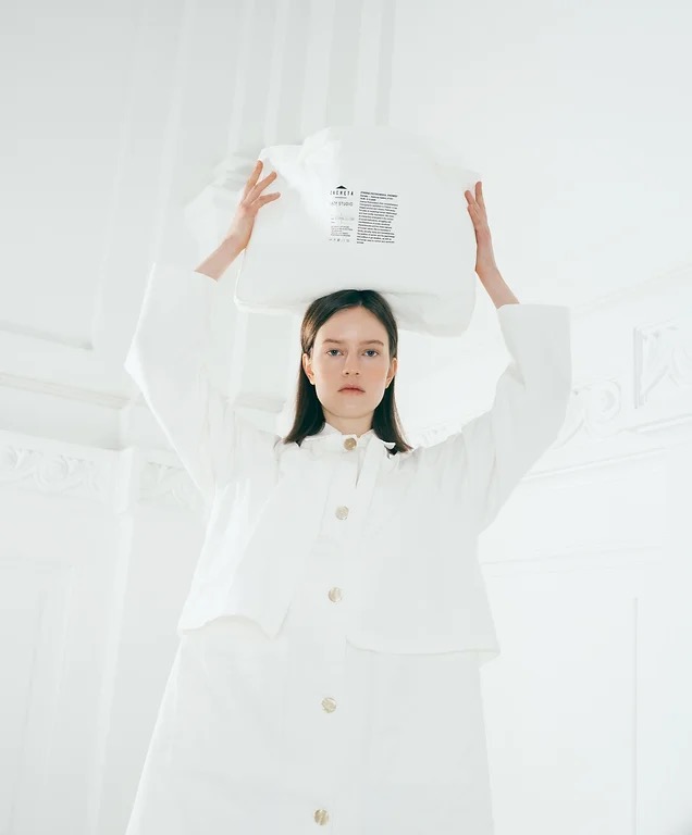Modelka w białym płaszczu z dużymi złotymi guzikami, trzyma nad głową dużą, białą torbę z nadrukiem, na jasnym, minimalistycznym tle.