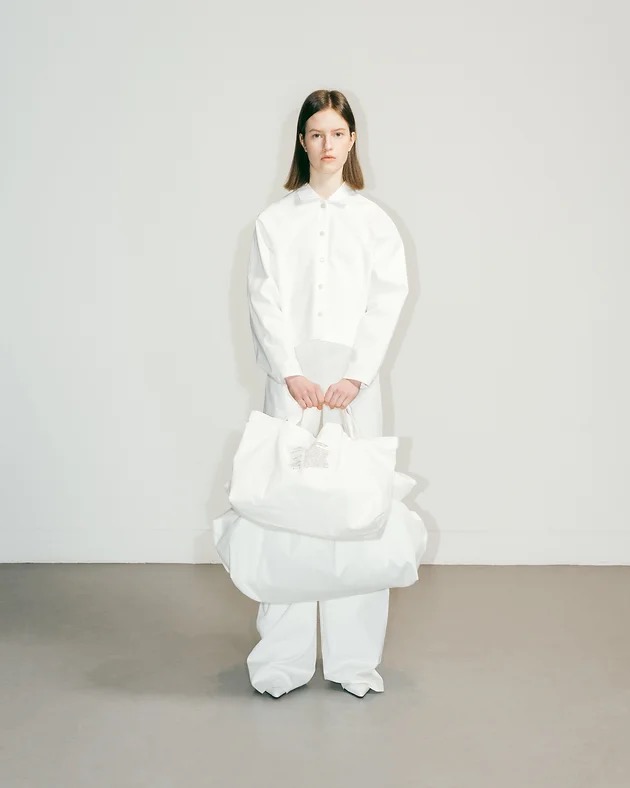 Modelka w białym komplecie (koszula i spodnie) trzymająca dwie duże, białe torby z materiału, na jednej z nich widoczna metka.