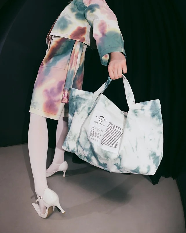 Modelka prezentuje komplet odzieży i torbę typu tote bag w stylu tie-dye, uzupełnione białymi rajstopami i lakierowanymi czółenkami.