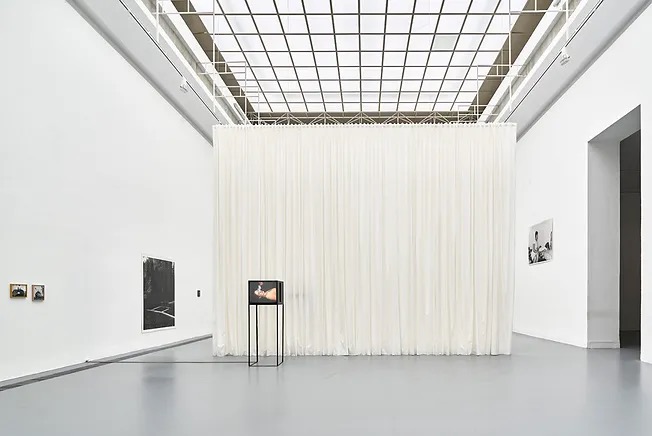 Ekspozycja w galerii sztuki: biała, drapowana tkanina oddziela przestrzeń, na pierwszym planie czarny stelaż z monitorem wyświetlającym wideo, na ścianach wiszą zdjęcia w ramkach.