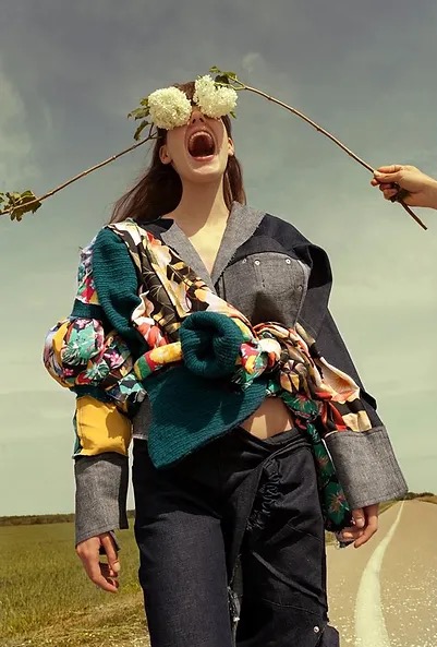 Awangardowa stylizacja z patchworkową kurtką, kwiatami zasłaniającymi oczy modelki i otwartymi ustami, na tle polnej drogi.