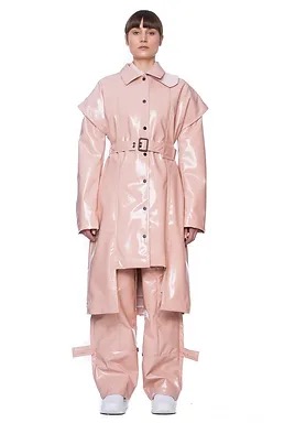 Modelka w błyszczącym, różowym płaszczu z paskiem i spodniach z paskami na nogawkach, na białym tle.
