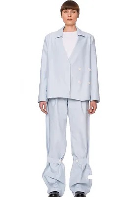 Modelka prezentuje komplet odzieżowy: luźny, jasnoniebieski blezer z białymi guzikami i szerokie spodnie z paskami na dole, na białym tle.