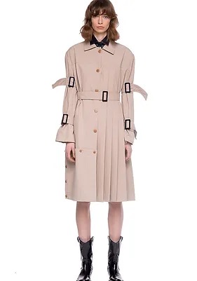 Modelka prezentuje beżowy trencz z plisowaną spódnicą, czarnymi klamrami na rękawach i pasku, oraz czarne lakierowane kowbojki na białym tle.