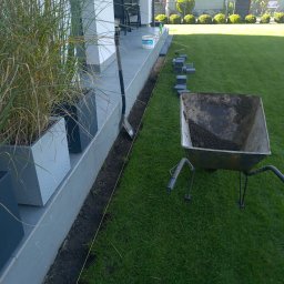 GK SYSTEM - Przygotowanie do układania obrzeży trawnikowych: wykopany rów, sznurek wyznaczający linię, kostki obrzeżowe ułożone wzdłuż ścieżki, taczka z ziemią i szpadel.