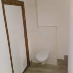 Wykończona toaleta z podwieszaną muszlą, drewnopodobne płytki na podłodze i drewniana rama drzwiowa.