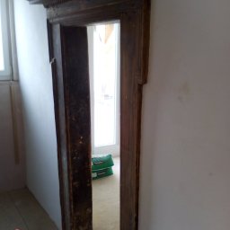 GK SYSTEM - Stara, drewniana framuga drzwiowa z dekoracyjnymi elementami, widoczne worki z zaprawą w tle, ściany w trakcie przygotowania do malowania.