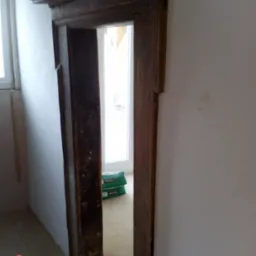 Stara, drewniana framuga drzwiowa z dekoracyjnymi elementami, widoczne worki z zaprawą w tle, ściany w trakcie przygotowania do malowania.