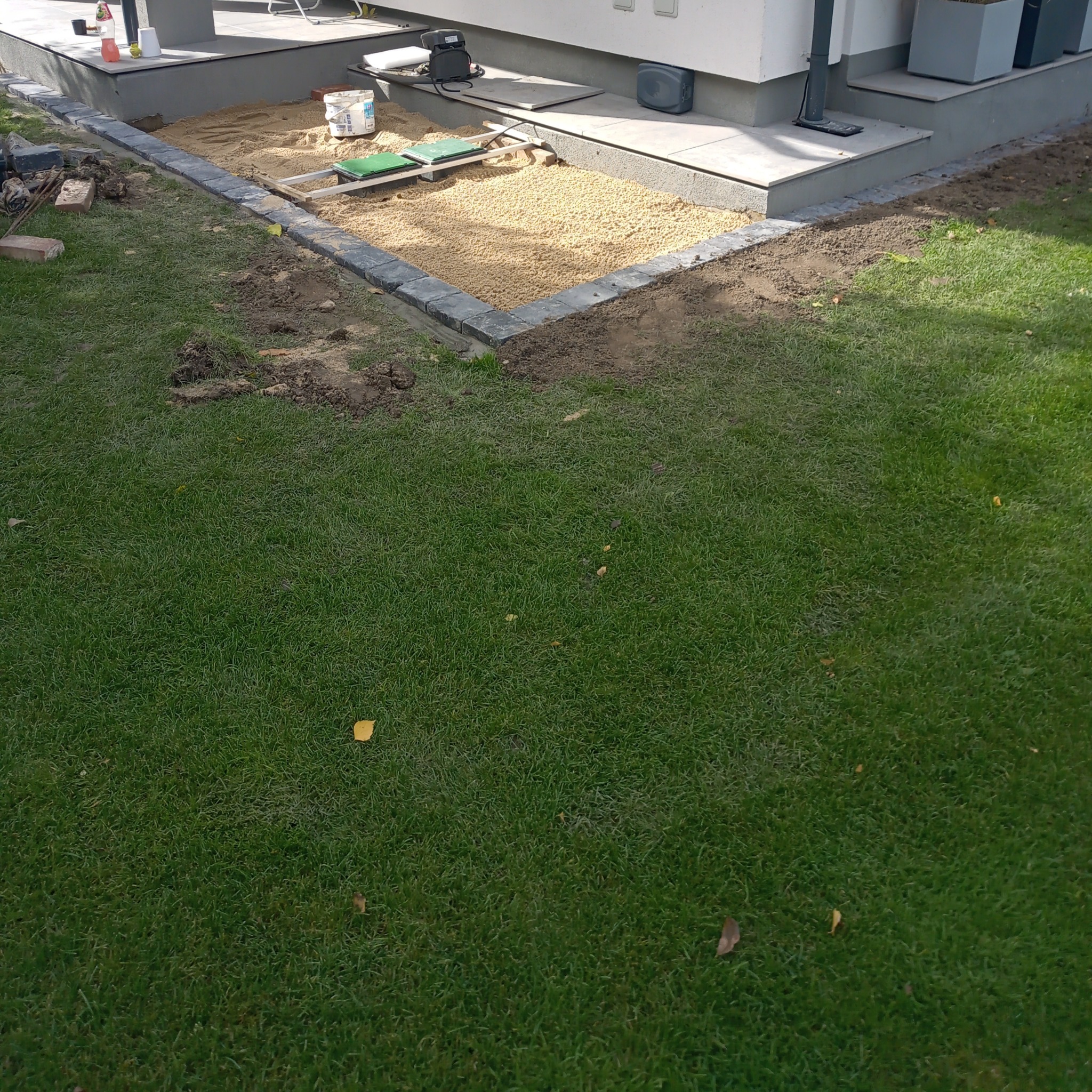 Przygotowanie podłoża z kamyczków pod taras, obrzeża z szarych kostek, trawnik w tle.