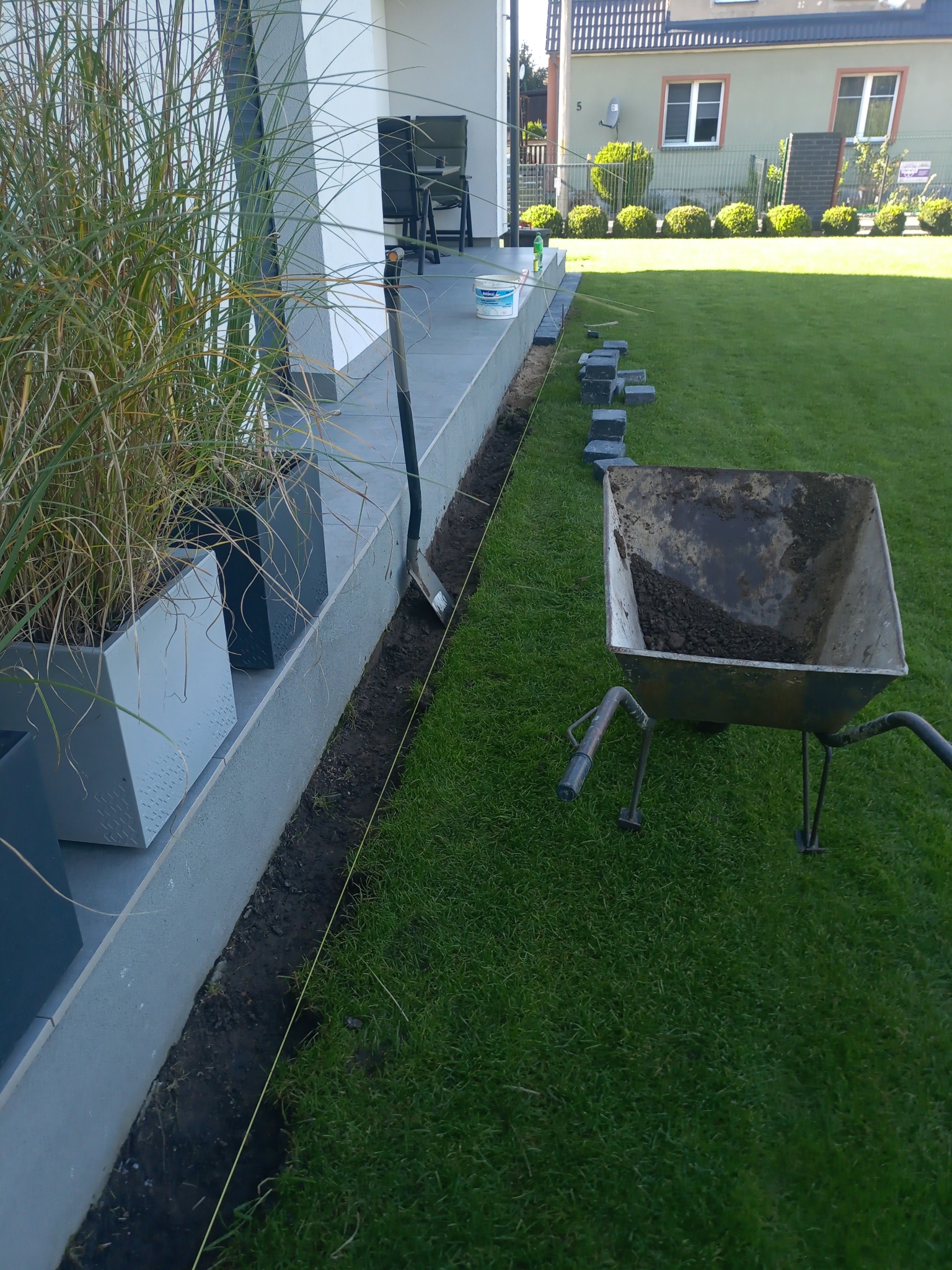 Przygotowanie do układania obrzeży trawnikowych: wykopany rów, sznurek wyznaczający linię, kostki obrzeżowe ułożone wzdłuż ścieżki, taczka z ziemią i szpadel.