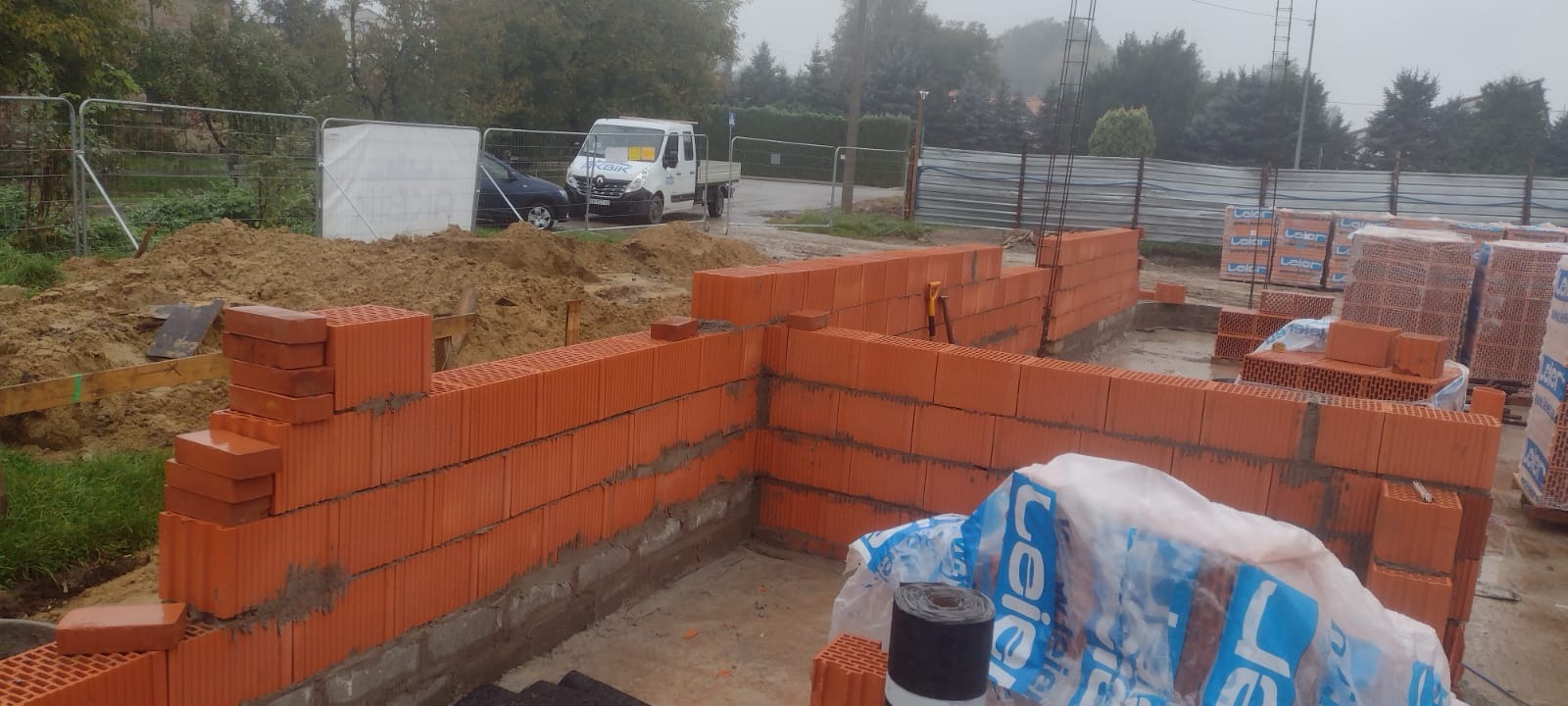 Wznoszenie ścian fundamentowych z czerwonej ceramiki poryzowanej na placu budowy, widoczny zapas bloczków, częściowo zabezpieczony folią, oraz firmowy samochód dostawczy w tle.