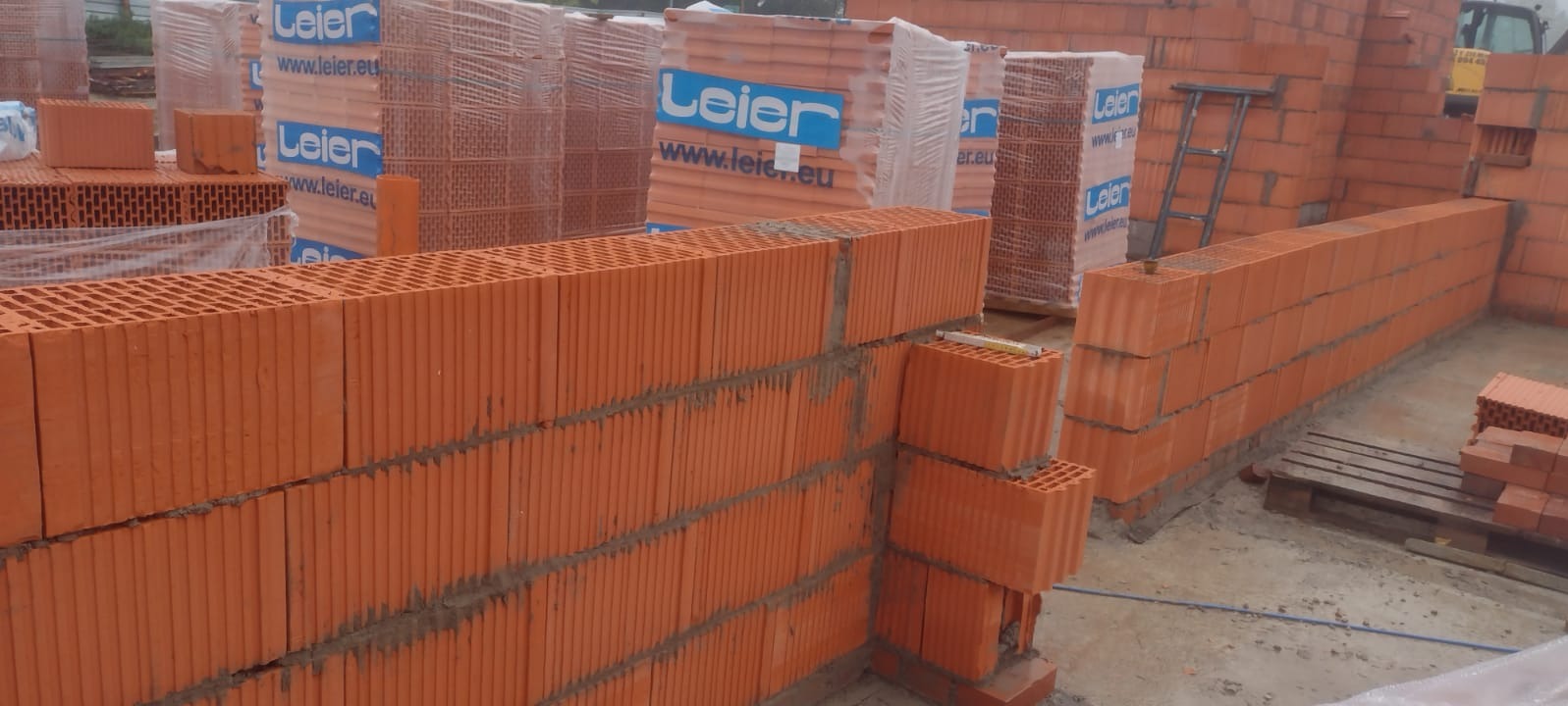 Wznoszenie muru z czerwonej ceramiki poryzowanej, widoczne spoiwo między warstwami, palety z zapakowanymi bloczkami w tle, drabina oparta o ścianę.