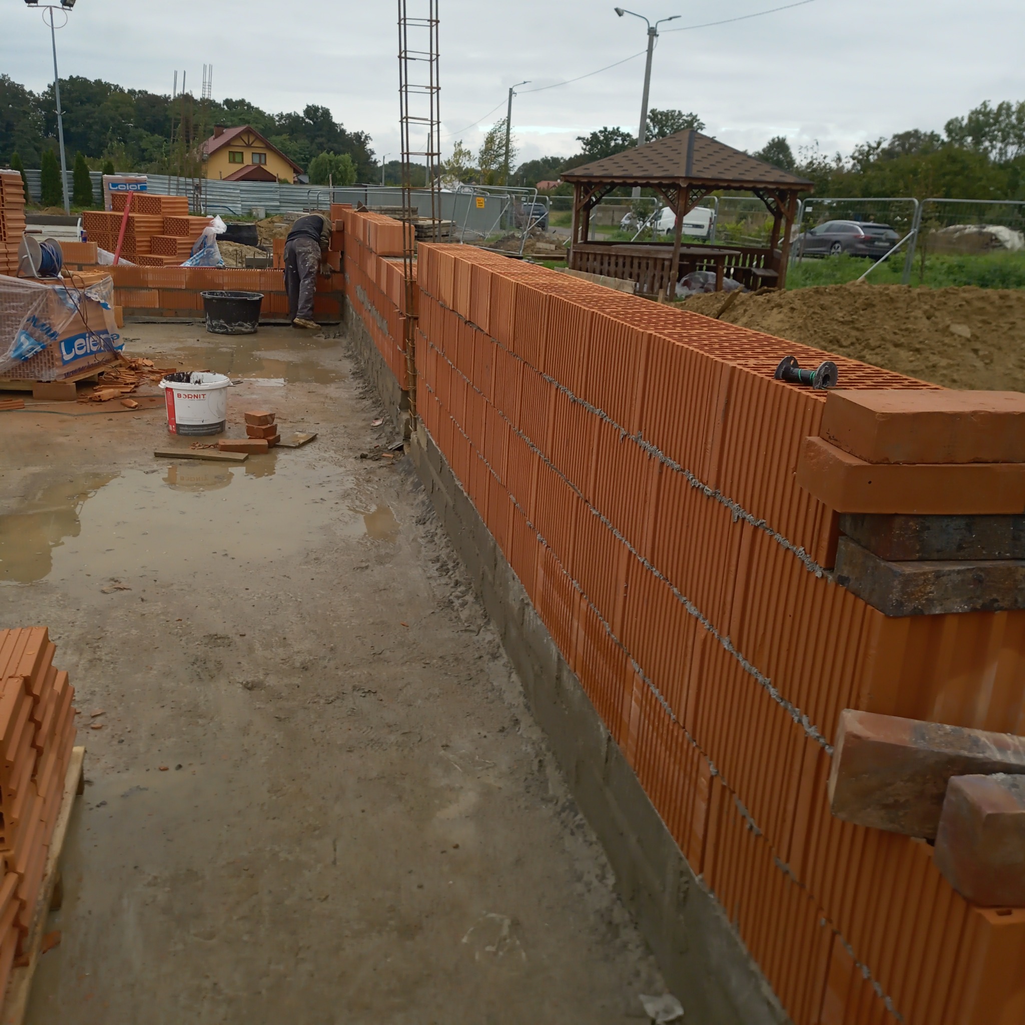 Budowa ściany z bloczków ceramicznych, widoczny pracownik murujący, paleta bloczków Leier oraz wiadro z zaprawą na mokrym podłożu placu budowy, w tle altana ogrodowa.