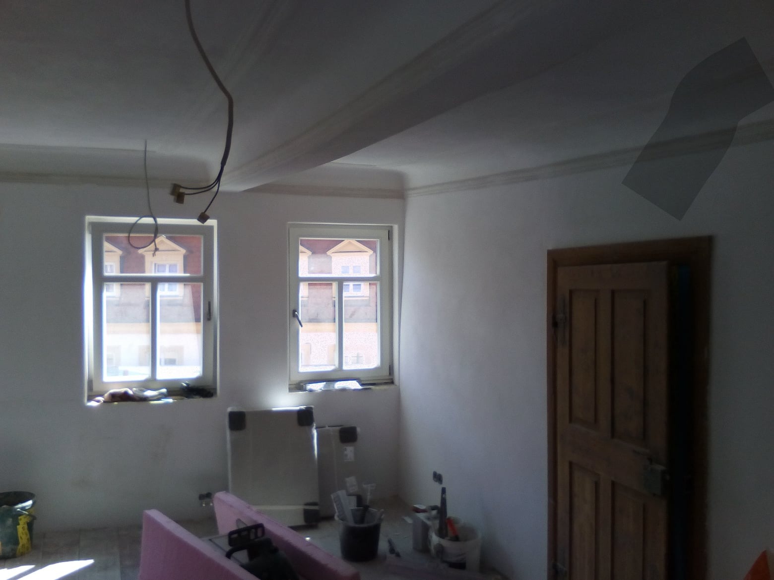 Pokój w trakcie prac wykończeniowych: surowe ściany, dwa okna z widokiem na budynki, widoczne narzędzia malarskie i wiązka kabli zwisająca z sufitu.