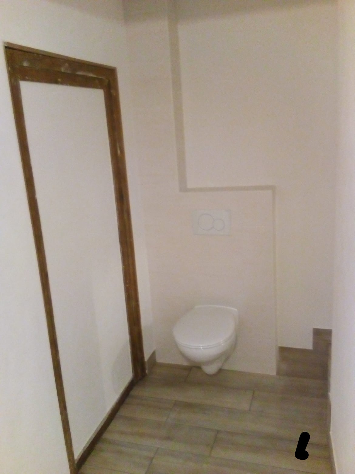 Wykończona toaleta z podwieszaną muszlą, drewnopodobne płytki na podłodze i drewniana rama drzwiowa.