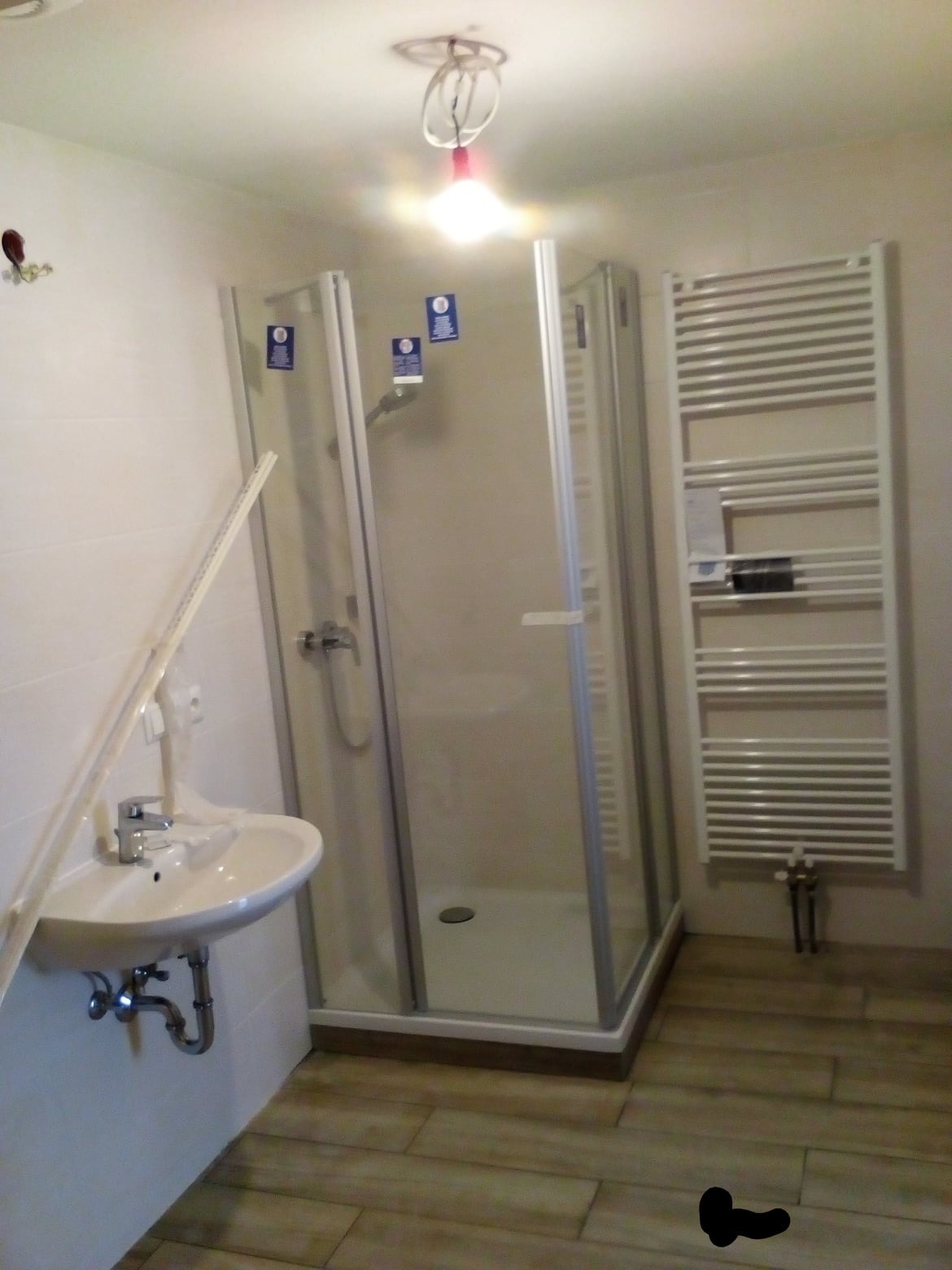 Nowoczesna łazienka z kabiną prysznicową, umywalką i grzejnikiem drabinkowym, widoczne elementy instalacji elektrycznej na suficie, płytki imitujące drewno na podłodze.