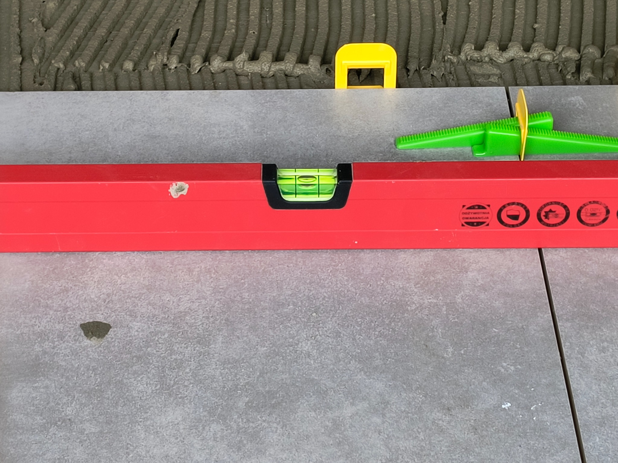 Poziomica na świeżo ułożonych, szarych płytkach, z widocznymi żółtymi i zielonymi klinami do poziomowania i fuga z klejem.