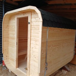 Cezary Gerek - Drewniana beczka sauny z otwartymi drzwiami, widoczne deski wewnątrz, dach pokryty czarnym gontem bitumicznym, całość zabezpieczona pasami transportowymi.