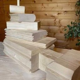 Cezary Gerek - Elementy konstrukcyjne z jasnego drewna ułożone warstwowo, prawdopodobnie do budowy domu z bali lub sauny, widoczne detale połączeń i obróbki drewna.