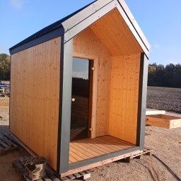 Cezary Gerek - Prosty, drewniany domek z ciemną ramą i szklanymi drzwiami, umieszczony na paletach na piaszczystym terenie, z widocznym polem i drzewami w tle.