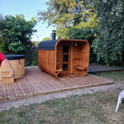 Cezary Gerek - Drewniana beczka z hydromasażem i beczkowa sauna z kominem na tarasie z desek, otoczone zielenią.