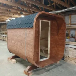 Gotowa beczka sauny z drewnianą konstrukcją i czarnym dachem z gontu bitumicznego, umieszczona w hali produkcyjnej na drewnianych podporach.
