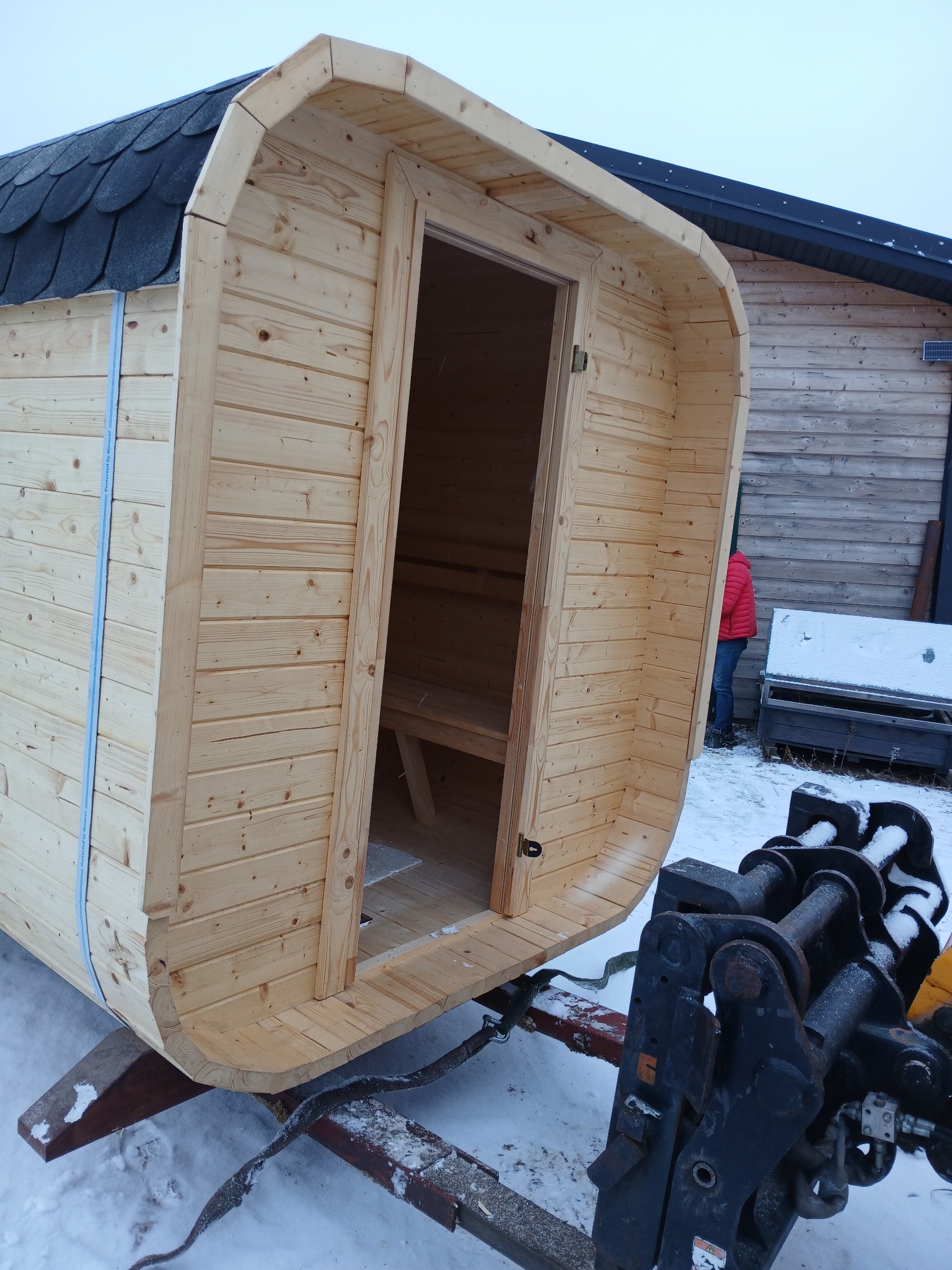 Przenośna, beczkowa sauna z jasnego drewna na przyczepie, widoczna w Pułtusku na tle zaśnieżonego terenu i budynku. Drzwi sauny otwarte, ukazują wnętrze z ławkami.