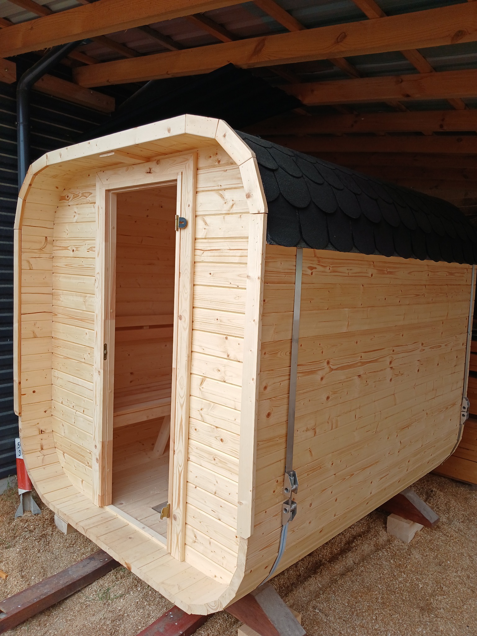 Drewniana beczka sauny z otwartymi drzwiami, widoczne deski wewnątrz, dach pokryty czarnym gontem bitumicznym, całość zabezpieczona pasami transportowymi.