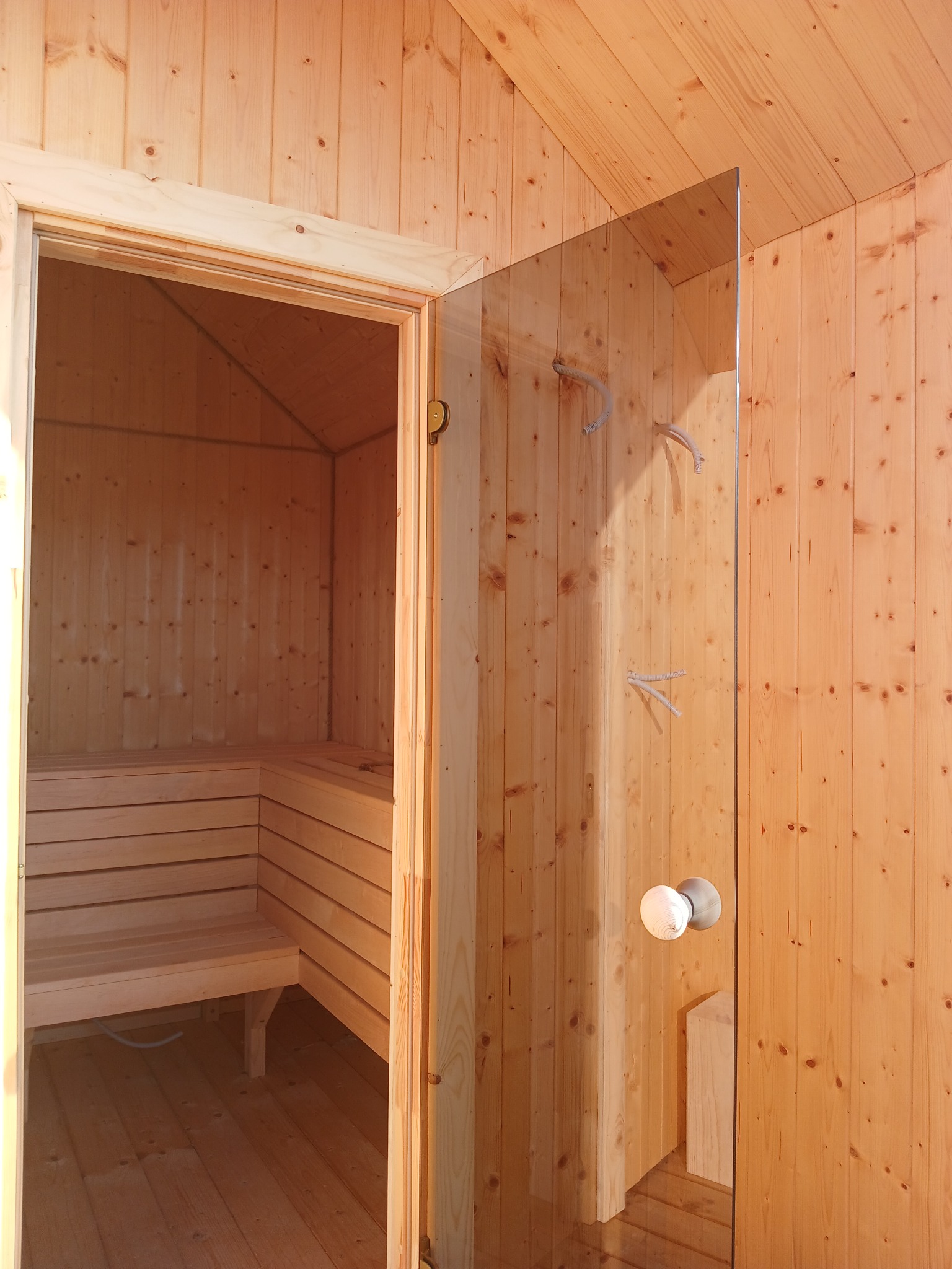Wnętrze sauny z drewnianymi ławkami widoczne przez otwarte, szklane drzwi z okrągłą klamką i wieszakami na ręczniki.
